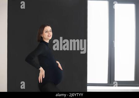 Giovane donna incinta in piedi su sfondo nero guardando la fotocamera a contatto con la sua pancia indossando abiti neri Foto Stock