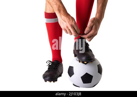 Sezione bassa del giocatore caucasico maschio giovane che lega la scarpa sulla palla di calcio su sfondo bianco Foto Stock