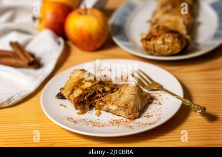 Un rotolo di strudel di torta di mele fresco cotto in casa con una fetta appetitosa di torta di mele sul piatto e mele fresche e bastoncini di cannella su un legno Foto Stock