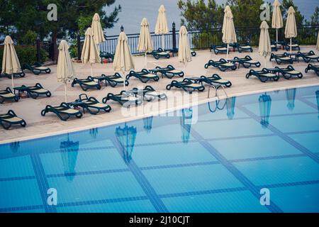 Sedie a sdraio in plastica e ombrelloni per la protezione del sole sulle piastrelle vicino alla piscina nella zona ricreativa Foto Stock