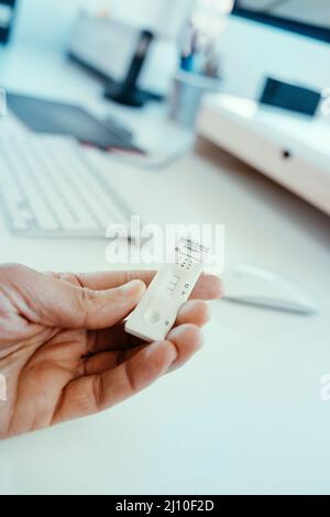 primo piano di un uomo in ufficio con un covid-19 antigene diagnostico dispositivo di prova con un risultato positivo in mano Foto Stock