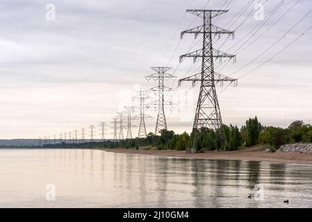 Piloni alti che supportano linee elettriche ad alta tensione sulla riva di un lago in una nuvolosa giornata autunnale Foto Stock