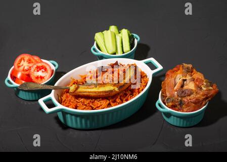 Jollof riso con banana fritta, pollo al forno. Riso con pomodori, cipolle, spezie. Cucina tradizionale nigeriana nazionale. Sfondo scuro. Foto Stock