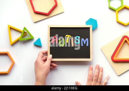 Testo autismo, parola sulla lavagna in colori arcobaleno. Triangoli e esagoni in legno annidato in vari colori audaci. Bordo gesso in mano umana. Foto Stock