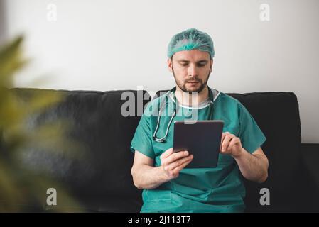 Medico che guarda il tablet nella stanza di riposo, vestendo abito chirurgico Foto Stock