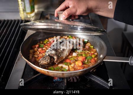 trota in padella, bollente in salsa di verdure, la mano di un cuoco solleva il coperchio. Foto Stock