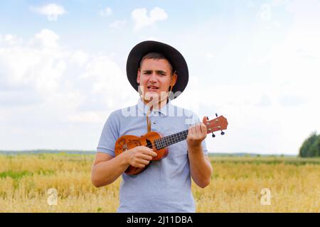 Concerto dal vivo. Giovane bel caucasico uomo giocare ukulele fuori. Il bel ragazzo hipster suona la chitarra ukulele su sfondo naturale. Maschio in un cowboy Foto Stock