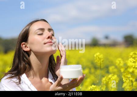 Donna con occhi chiusi che applica crema idratante sulla guancia una giornata di sole Foto Stock