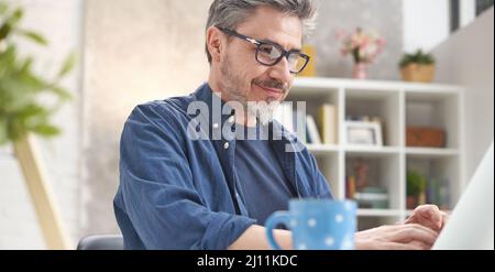 Uomo felice con il computer portatile a casa. Uomo d'affari che lavora alla scrivania in ufficio. Ritratto di età matura, età media, uomo adulto medio, barbuto, occhiali, look sorridente e autentico. Foto Stock