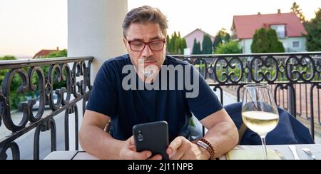 Ritratto all'aperto di uomo medio adulto nel 50s, felice sorriso sicuro con il telefono. Posti a sedere sulla terrazza del ristorante. Età matura, età media, barbata. Foto Stock