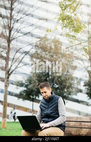 Uomo libero in abiti casual, utilizzando un computer portatile, edificio moderno sullo sfondo. Lavorare in remoto in città Foto Stock