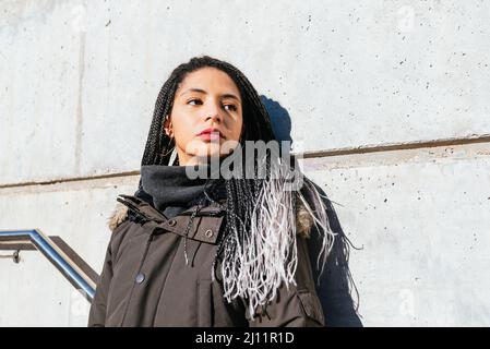 Donna tranquilla con lunghi capelli intrecciati e in abiti caldi in piedi vicino alla parete in strada e guardando via Foto Stock