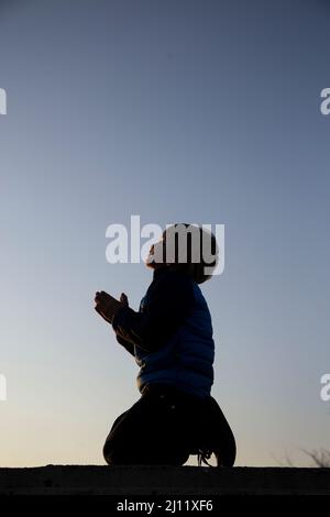 Silhouette di un ragazzo inginocchiato, piegato le mani in preghiera, un appello a Dio. Bambini contro la guerra. Un bambino ucraino chiede pace per la sua patria Regno Unito Foto Stock