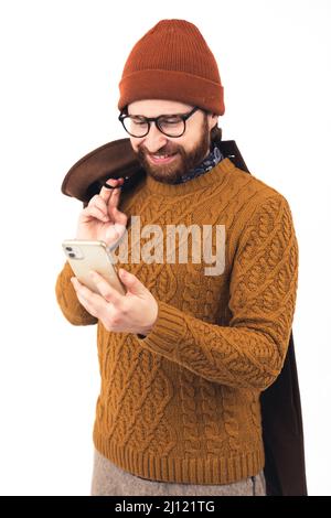 Verticale medio scatto di un giovane bel sorridente uomo caucasico che guarda il suo smartphone e sorridente cappotto di tenuta sulla spalla abbigliamento casual marrone sfumature di colori vestiti bianco sfondo studio shot . Foto di alta qualità Foto Stock