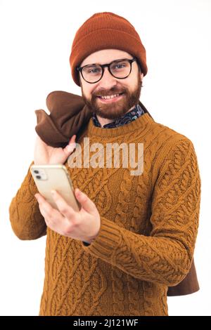 Ritratto di un giovane uomo caucasico sorridente sorridente che tiene smartphone in una mano e cappotto sulla spalla bianco sfondo studio scatto utilizzando il concetto di connessione smartphone. Foto di alta qualità Foto Stock