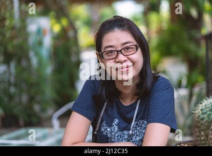 Ritratto di donna asiatica di mezza età indossa occhiali, acconciatura nera corta, seduta in un giardino, volto sorridente, spazio per il testo. Foto Stock