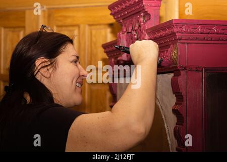 Donna caucasica sorridente colorazione legno ornato armadio in rosso con grande pennello con porte sullo sfondo. Riutilizzo di oggetti antichi. Casa Foto Stock