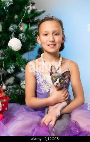 Ragazza sorridente in abito viola fantasia che tiene giocoso nazionale canadese Sphynx Cat seduta vicino albero di Natale la vigilia di Natale. Cute aspetto pre-adolescente ragazza Foto Stock