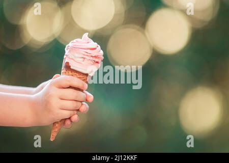 La mano del bambino che tiene un grande gelato alla fragola in un cono del waffle su uno sfondo bokeh Foto Stock