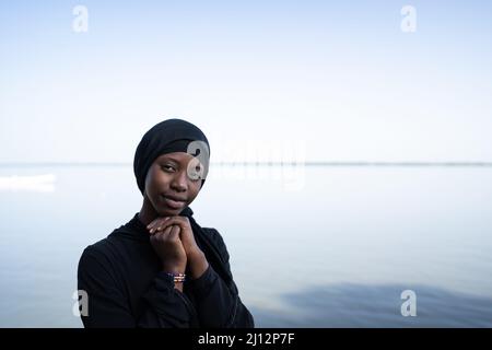 Bella giovane ragazza africana vestita di nero con le mani aggrappate sotto il mento, guardando tranquillamente la macchina fotografica; pace interiore della mente e spirituale Foto Stock