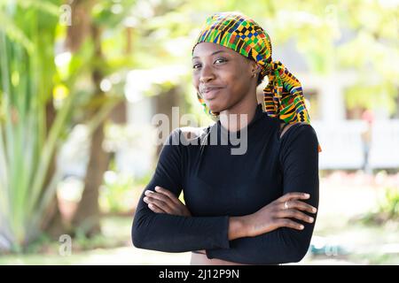 Giovane e intelligente bellezza africana che indossa una sciarpa colorata e annodata sulla testa e un maglione nero di dolcevita, sorridendo mischievamente alla macchina fotografica; simboliz Foto Stock