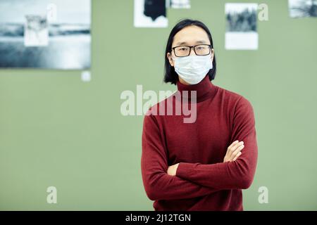 Giovane uomo asiatico che indossa occhiali e maschera protettiva sul viso in piedi con le braccia incrociate nella galleria d'arte moderna guardando la macchina fotografica Foto Stock