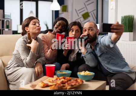 Gruppo di persone che prendono selfie e festeggiano con bevande in ufficio dopo l'orario di lavoro. L'uomo e le donne si divertono con le foto sullo smartphone, gustando bevande alcoliche e spuntini a festa. Foto Stock