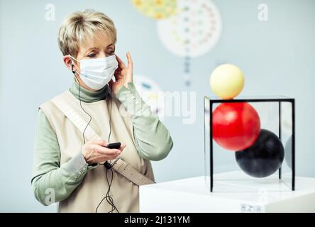 Ritratto di donna anziana con maschera protettiva sul viso con audioguida alla mostra nel museo d'arte contemporanea Foto Stock