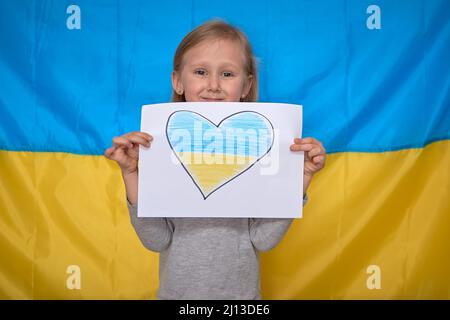 Le mani della ragazza tengono il banner di carta con il cuore disegnato giallo-blu sullo sfondo della bandiera Ucraina. I bambini sostengono l'Ucraina, arrestano la guerra, rimangono con hashtag Ucraina Foto Stock