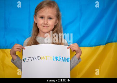 Le mani della ragazza tengono il banner di carta con tracciato giallo-blu staywithUcraina hashtag sullo sfondo della bandiera Ucraina. I bambini sostengono l'Ucraina, arrestano la guerra Foto Stock