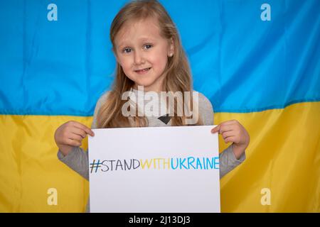 Le mani della ragazza tengono il banner di carta con tracciato giallo-blu staywithUcraina hashtag sullo sfondo della bandiera Ucraina. I bambini sostengono l'Ucraina, arrestano la guerra Foto Stock