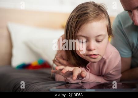 Padre e sua bambina con sindrome di Down sdraiati a letto e usando una tavoletta a casa. Foto Stock