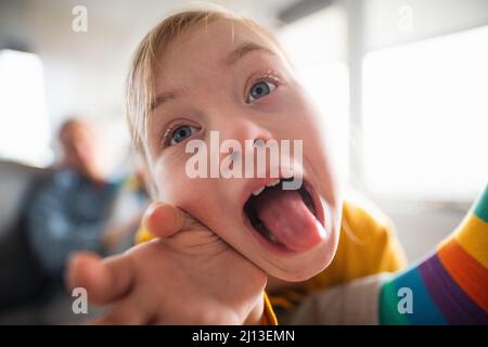 Primo piano di ragazza piccola allegra con la sindrome di Down che attacca la lingua fuori a casa. Foto Stock