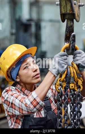 Giovane ingegnere femminile contemporaneo che appende un mazzo di catene metalliche su gancio di gru o altra moderna macchina industriale Foto Stock