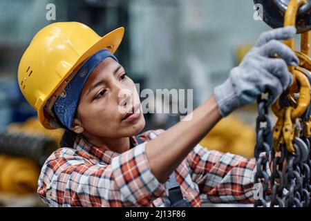 Giovane ingegnere ispanico femminile in hardHat e guanti di controllo catene di moderna macchina industriale durante il lavoro in magazzino Foto Stock