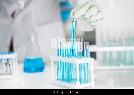 Mano guanto di chimico femminile o biotecnologo che prende uno dei molti matracci contenenti sostanza chimica liquida blu in laboratorio Foto Stock