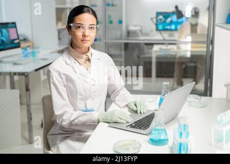 Giovane chimica femminile contemporanea con un computer portatile che inserisce nuovi dati scientifici mentre si siede sul posto di lavoro e si guarda la fotocamera Foto Stock