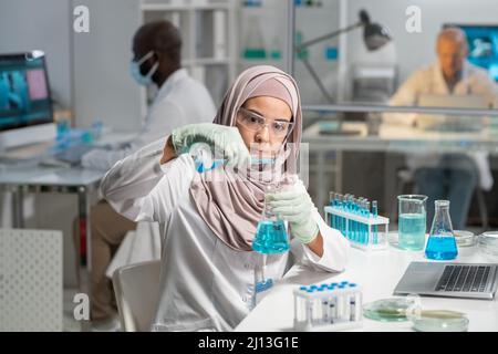 Giovane ricercatrice musulmana seria in hijab, guanti e occhiali protettivi versando sostanza liquida in fiasca con fluido blu Foto Stock