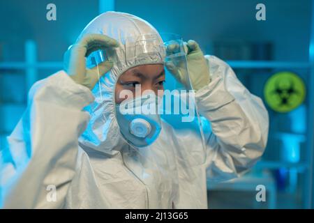 Giovane scienziata asiatica in tuta a rischio biologico, guanti e respiratore mettendo schermo protettivo sul viso di fronte al laboratorio Foto Stock