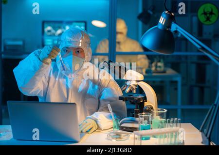 Giovane virologo femminile in tute protettive, respiratore, guanti e schermo facciale studiando campione di nuovo vaccino in laboratorio Foto Stock