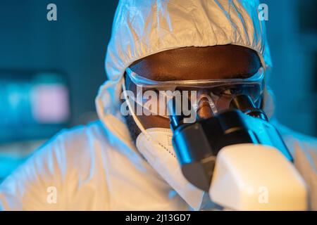 Primo piano del giovane scienziato afroamericano in tute protettive, respiratore e occhiali guardando al microscopio in laboratorio Foto Stock