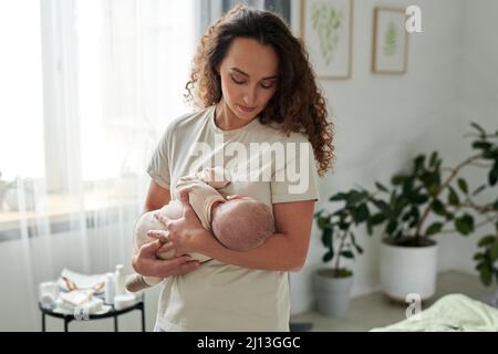 Giovane madre contemporanea in t-shirt che tiene il neonato dal petto mentre lulling lui in camera da letto a metà giornata Foto Stock