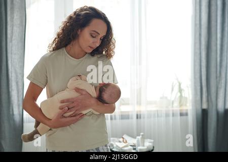 Giovane donna serena con il figlio accanto al petto lullandolo mentre si trova in camera da letto contro tende bianche e grigie Foto Stock