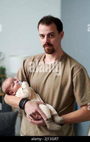 Giovane uomo in t-shirt casual che tiene il bambino ragazzo in abito rompro mentre si trova di fronte alla macchina fotografica e lulling bambino Foto Stock
