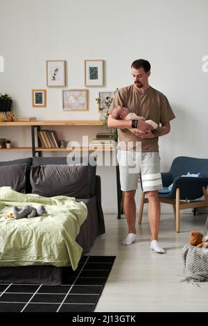 Giovane attento padre in casualwear che si muove lungo comodo letto matrimoniale con giocattoli mentre tiene il suo adorabile figlio del bambino sulle mani Foto Stock