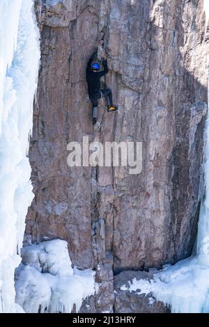 Un arrampicatore sportivo su una scalata mista, utilizzando assi di ghiaccio e ramponi per scalare una parete di roccia nel Parco di ghiaccio di Ouray in Colorado. La sezione di roccia è circondata Foto Stock