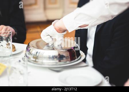 Processo di servire cena di lusso in un piatto di chiodi di garofano con un coperchio, cameriere che serve il pasto principale sotto piatto d'argento su un banchetto nel ristorante o Foto Stock