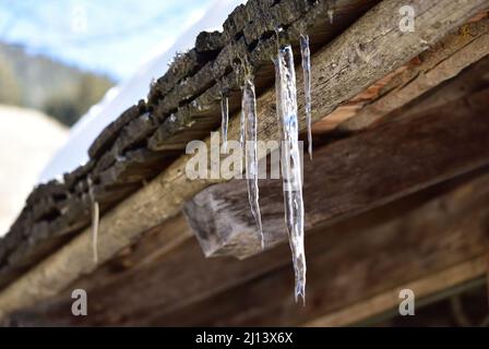 L'aria ancora fredda forma le ghiacciate mentre la neve si scioglie sul tetto della capanna Foto Stock