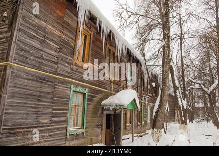 Le ciclicole sempre più trasparenti si appendono sul bordo del tetto. Sullo sfondo del muro di legno della vecchia casa. Grandi cascate, anche belle file. Nuvoloso giorno d'inverno, luce tenue. Foto Stock
