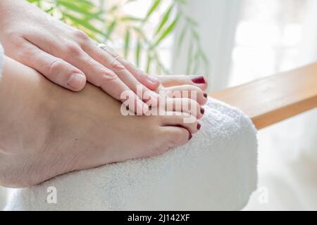 mani di una giovane donna che massaggiano i piedi con unghie dipinte di rosso dopo aver ottenuto un pedicure a casa Foto Stock
